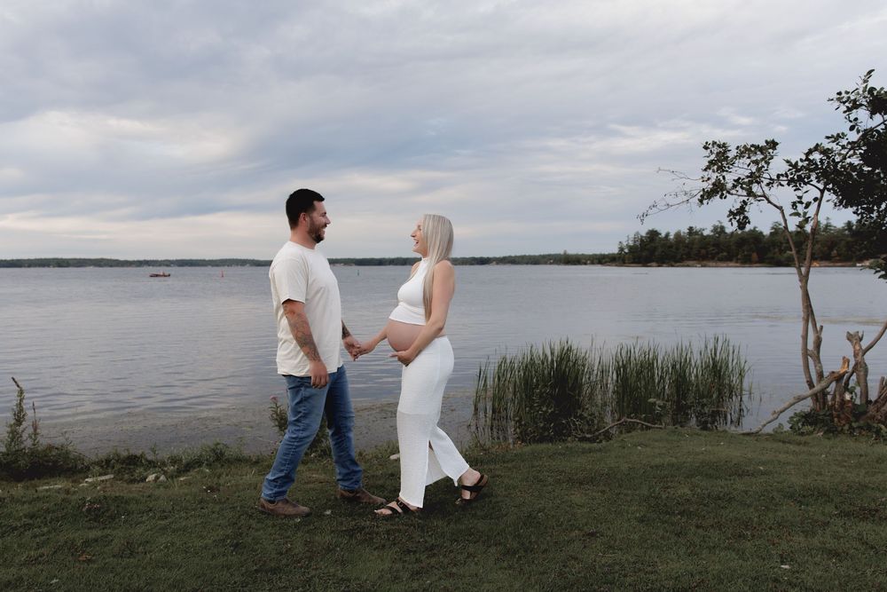 Two people sit together on a grassy shore overlooking a tranquil lake on an overcast day.