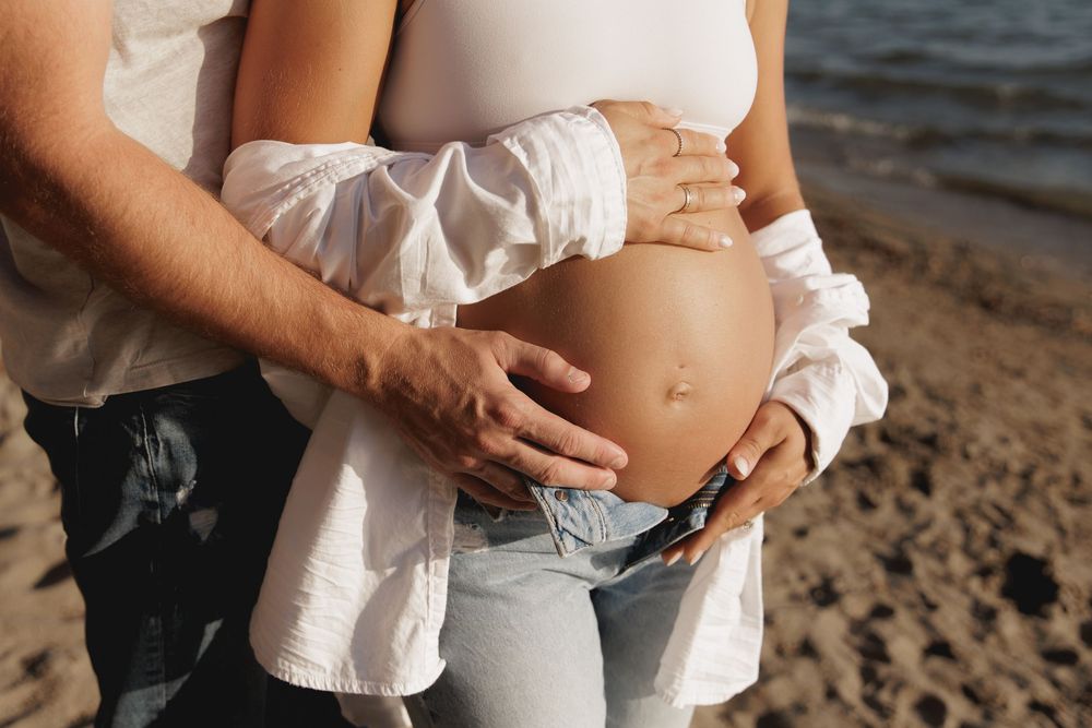 Hands tenderly cradle a pregnant belly on a sandy beach at sunset, wearing a white shirt and jeans.