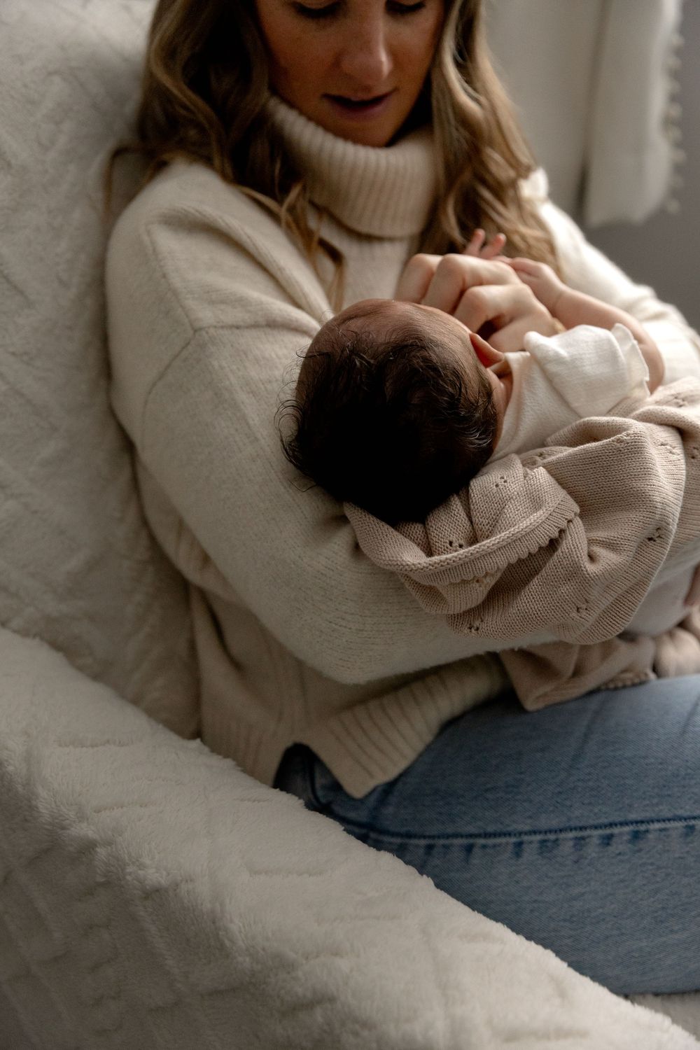 Parent in cozy cream sweater tenderly holds and nurtures sleeping newborn baby on blue surface.