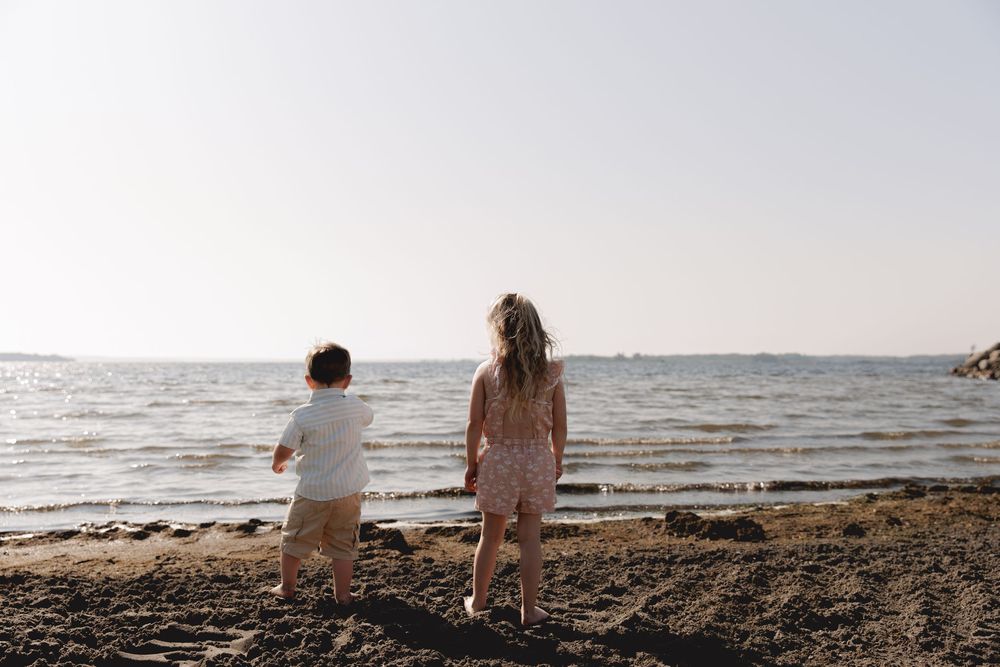 Two people stand together on a sandy beach looking out at the calm ocean during a bright sunny day.
