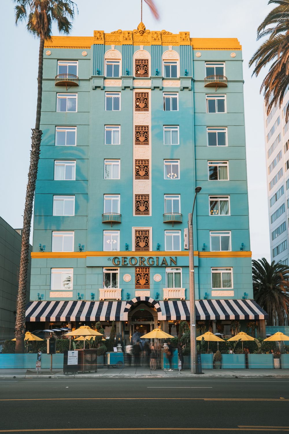 Art deco hotel building in turquoise with yellow umbrellas and striped awnings along a palm-lined street.