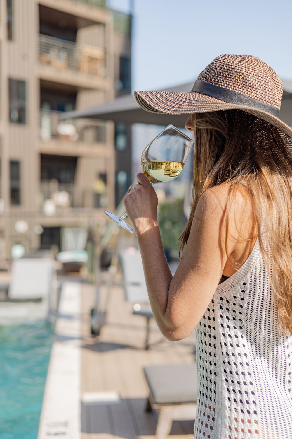 Person in straw hat and polka dot top sips wine by a resort pool on a sunny day.
