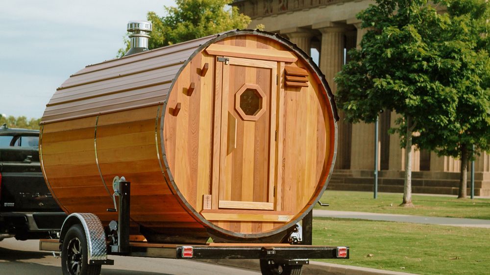 A wooden barrel-shaped sauna trailer with a circular door and copper-toned finish parked on grass near a building.