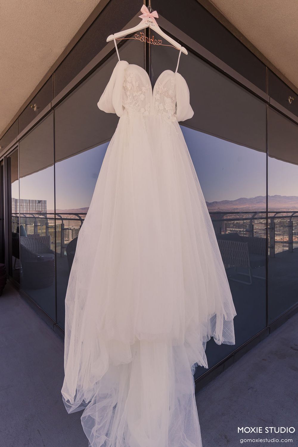 White wedding dress hanging on display against a modern glass window backdrop with mountain views at sunset.