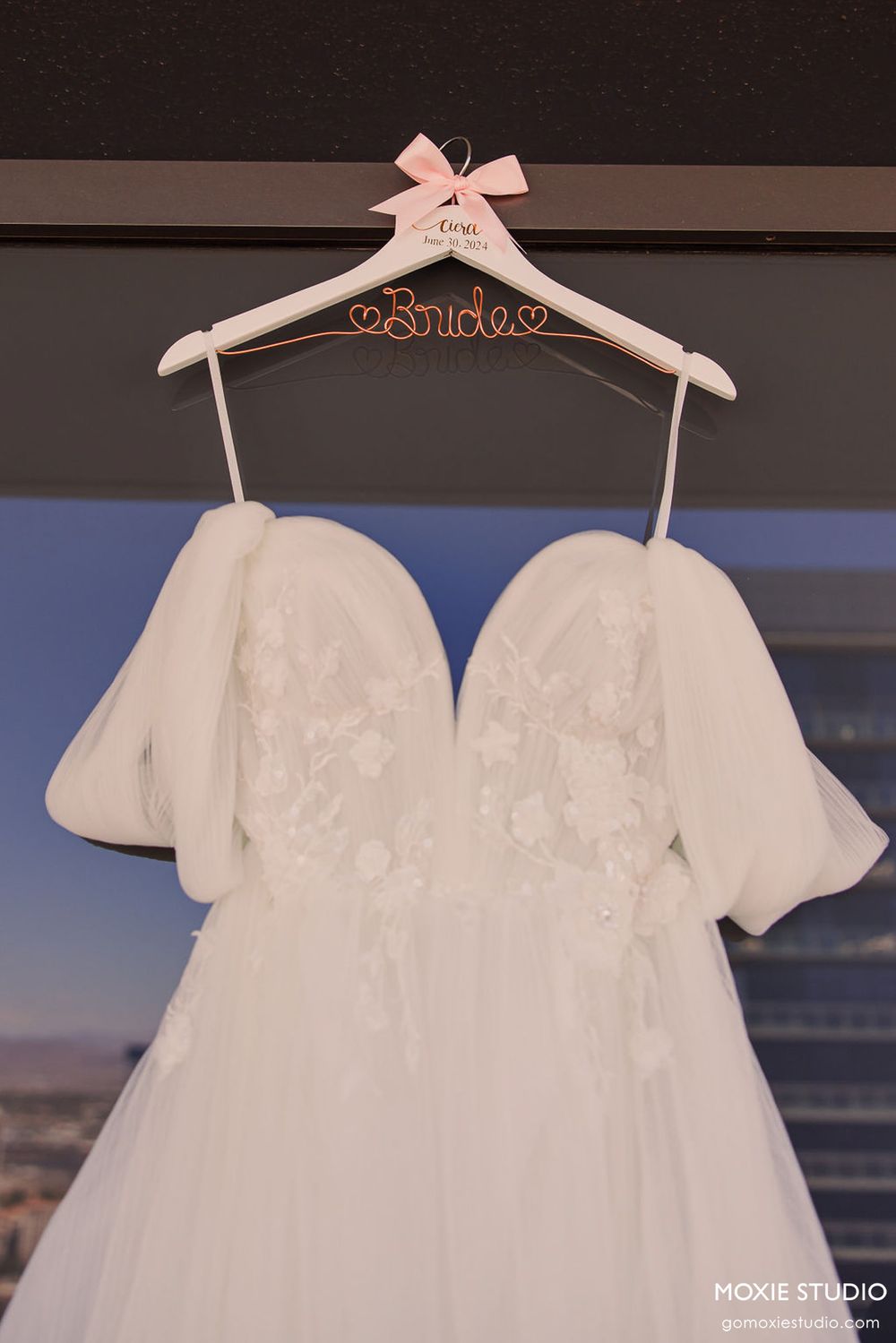 White baptismal or christening dress with delicate lace details displayed on a pink bow adorned hanger.