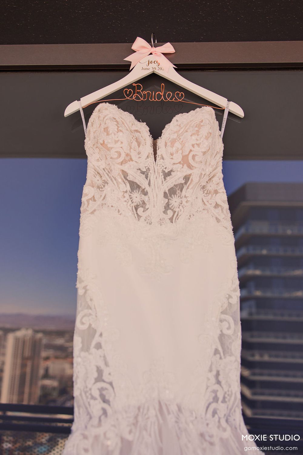 Elegant white lace wedding dress with intricate floral details displayed on a custom bridal hanger against a city skyline view.