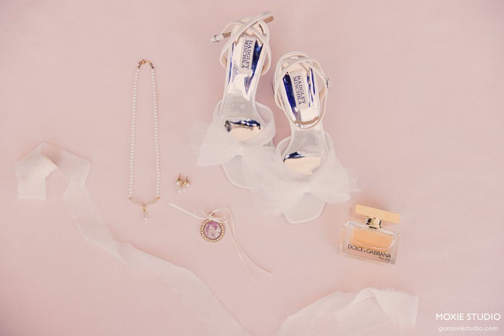 Delicate wedding accessories including jewelry, perfume bottles, and ribbons arranged on a soft pink surface.