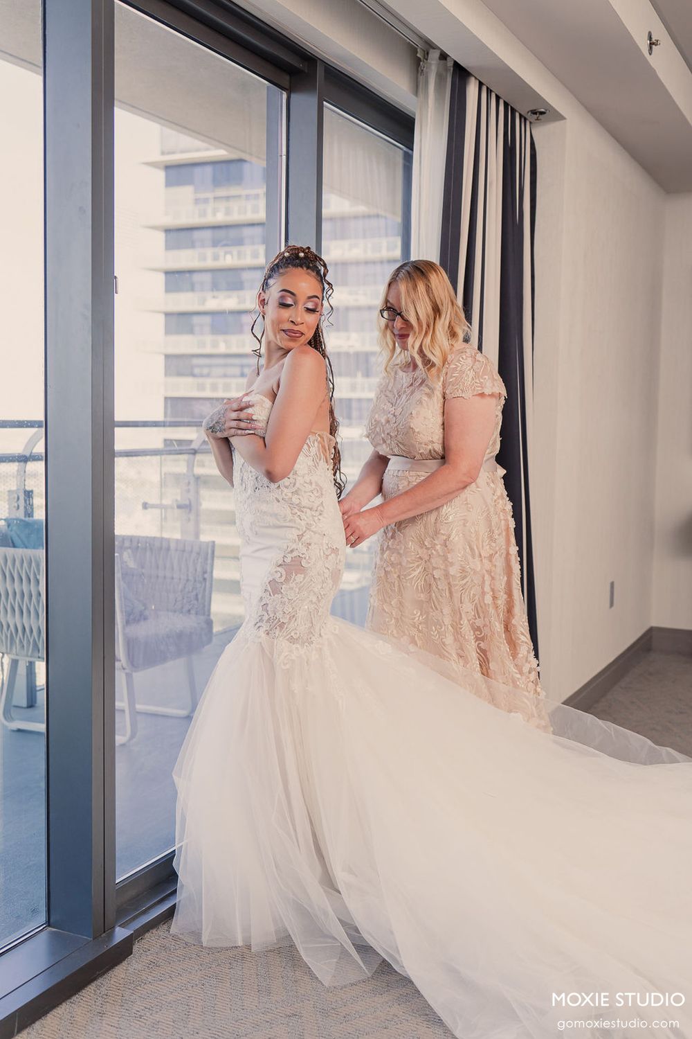Two people in elegant lace and tulle wedding gowns stand by large windows in a bright modern room.