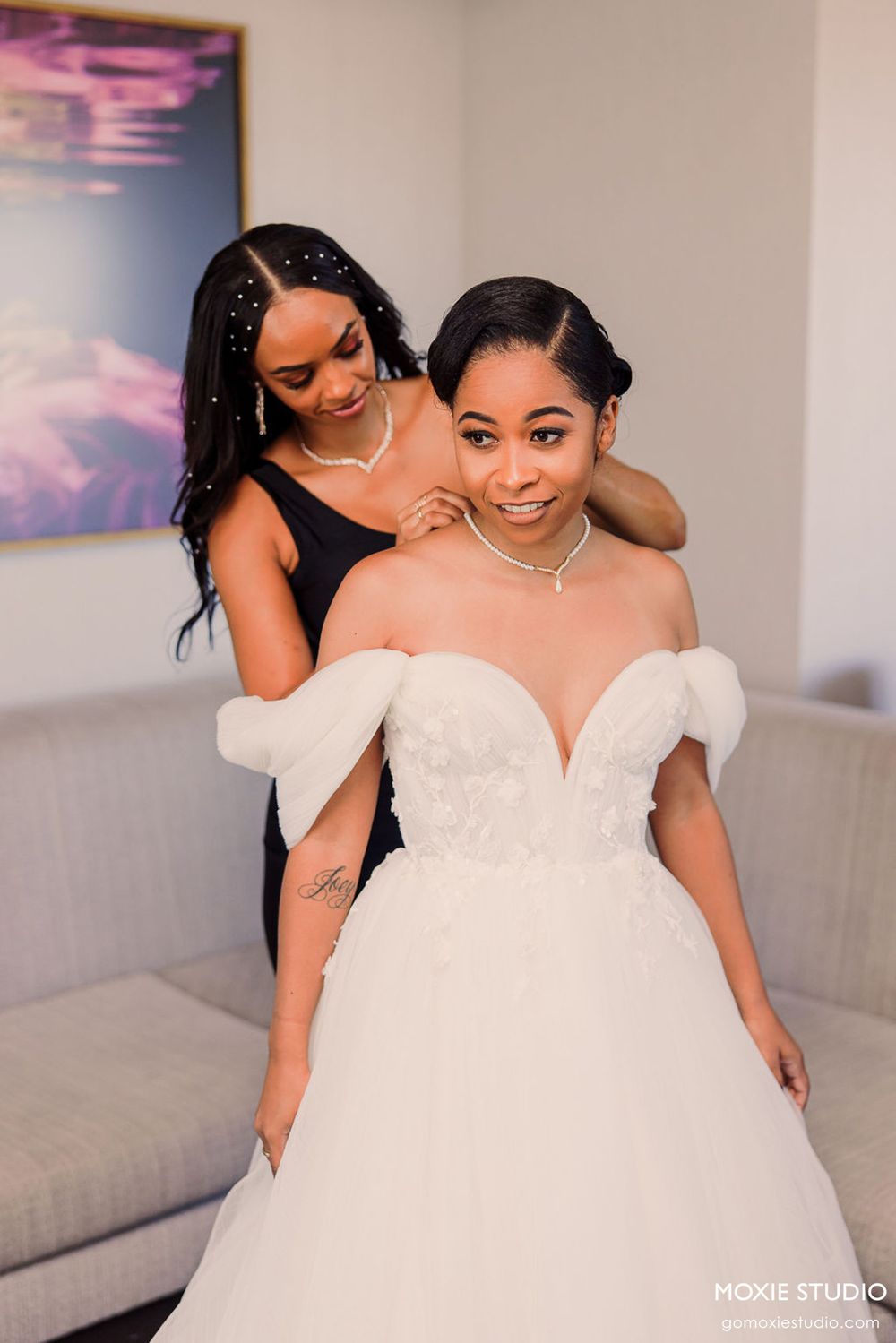 Bride in elegant white wedding gown shares intimate moment with bridesmaid in dark dress at indoor venue.