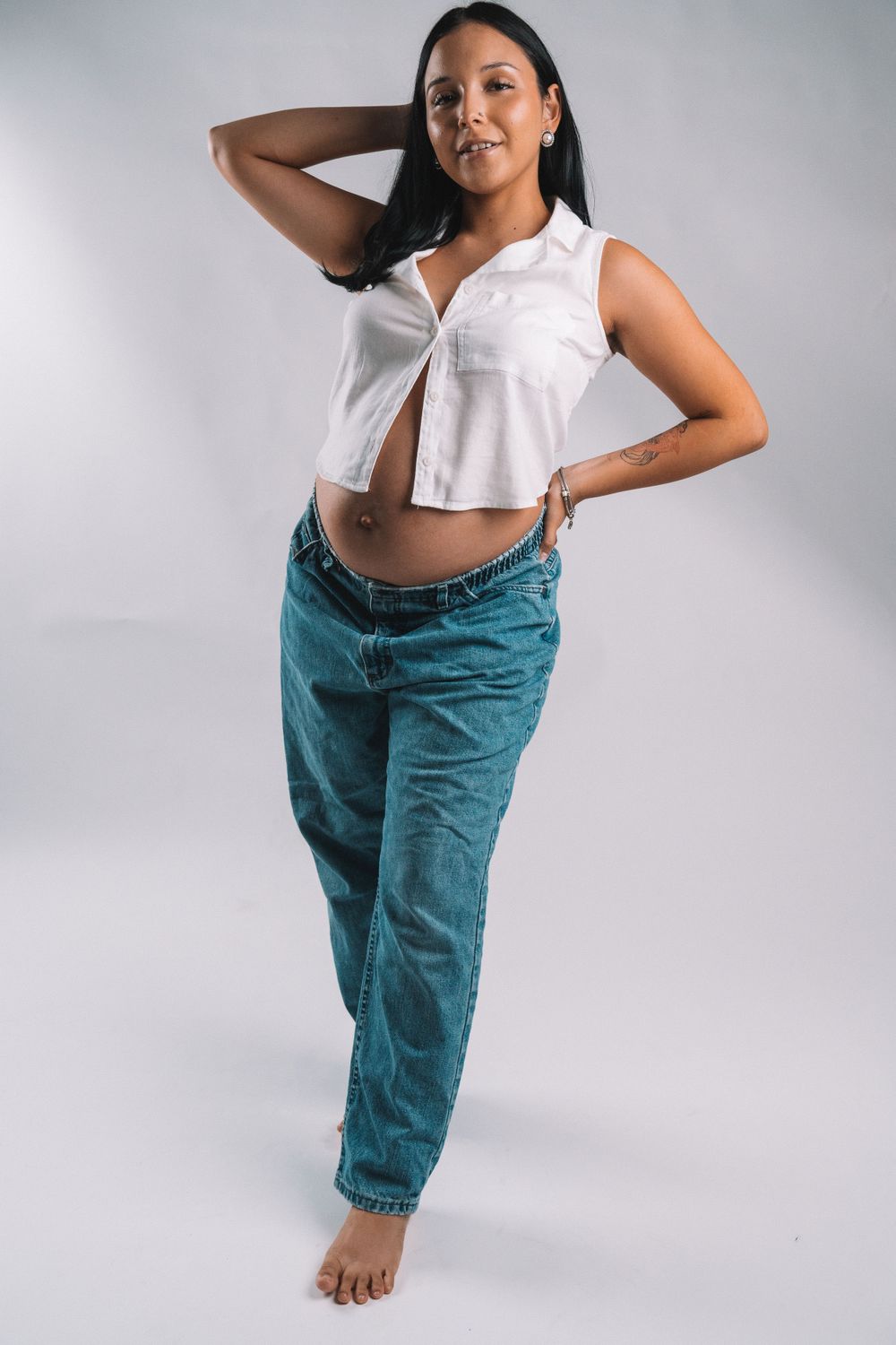 Woman in white sleeveless top and teal jeans poses against light background in casual fashion shoot.