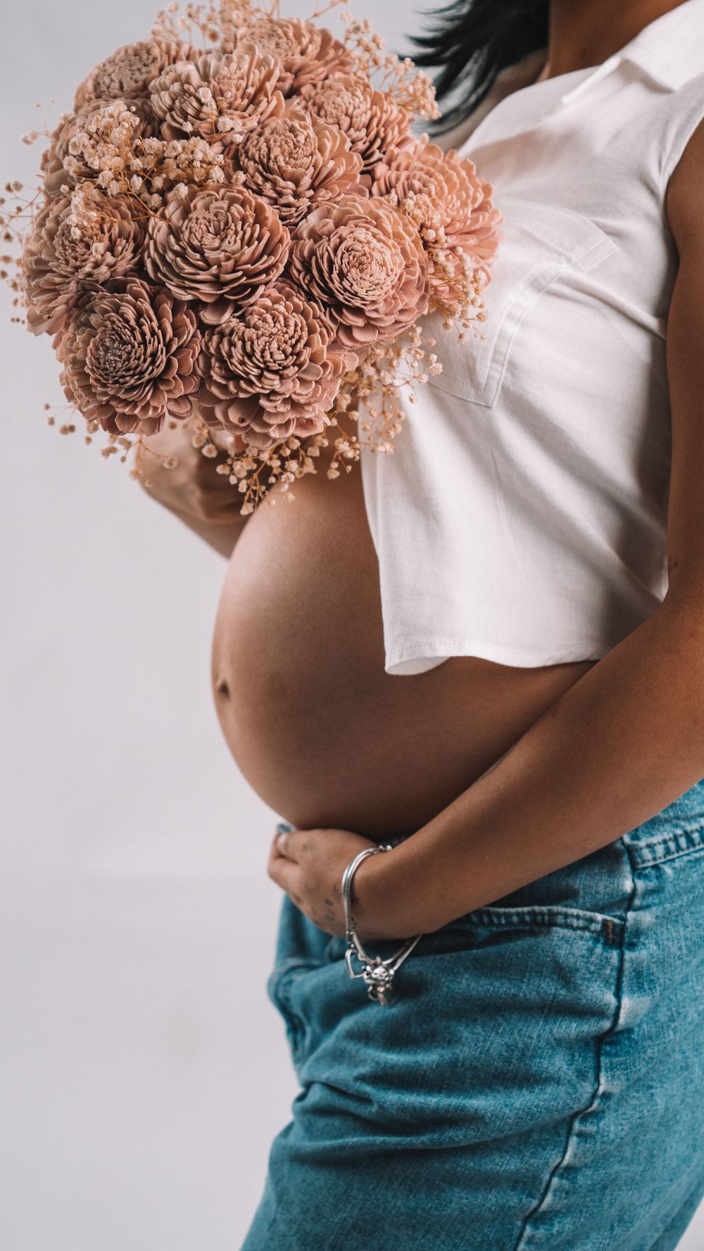 Close up of dried pink floral bouquet held against white top and denim in maternity photo.