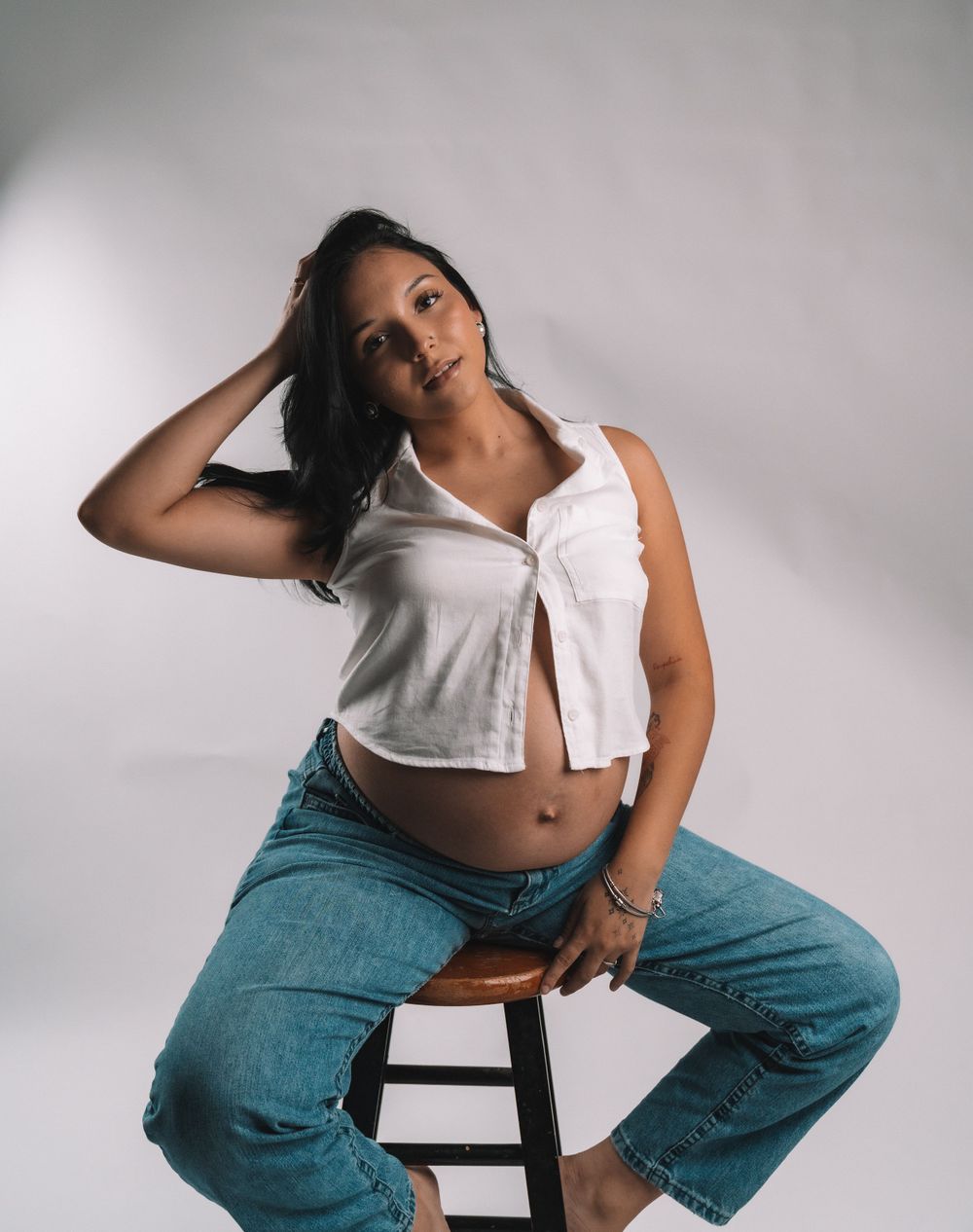 Casual portrait pose sitting on stool wearing white crop top and teal jeans.