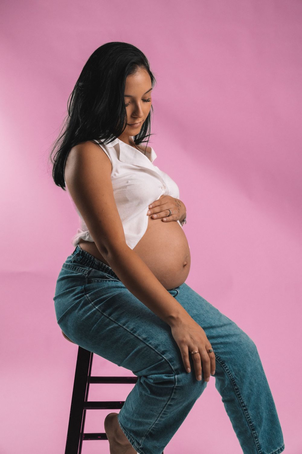 Maternity portrait against pink background showing profile pose on wooden stool.