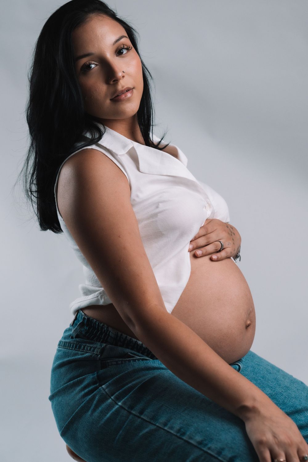 Side view maternity portrait against gray background in casual white top and jeans.