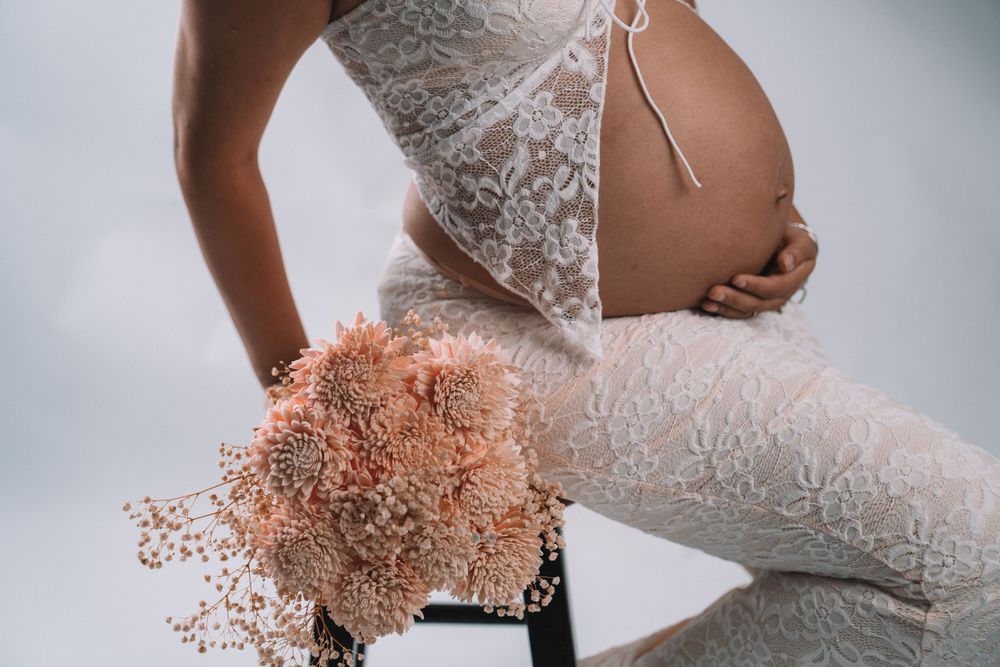 Elegant maternity photo featuring white lace dress and pink dried flowers against light background.