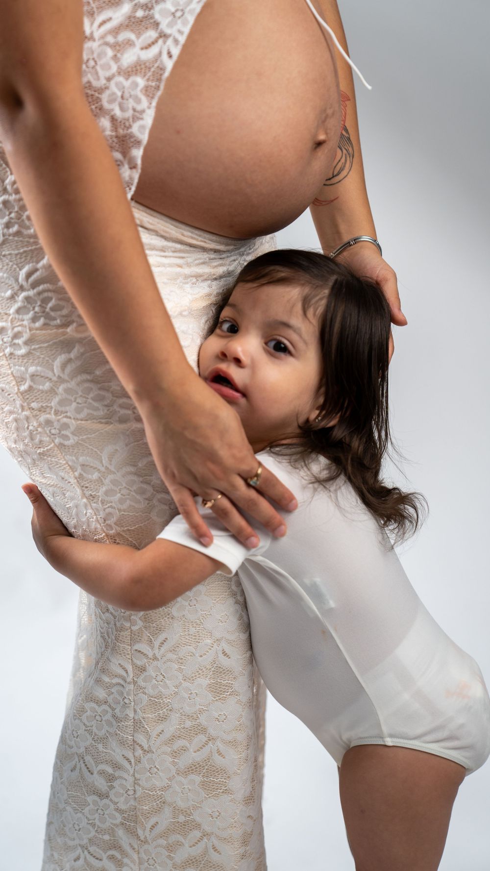 Sweet maternity moment with toddler hugging pregnant belly in white lace dress.