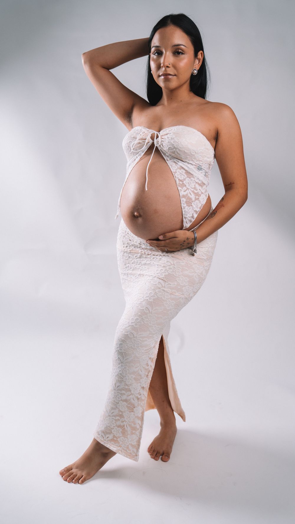 Artistic maternity portrait in floor-length white lace dress against gray backdrop.
