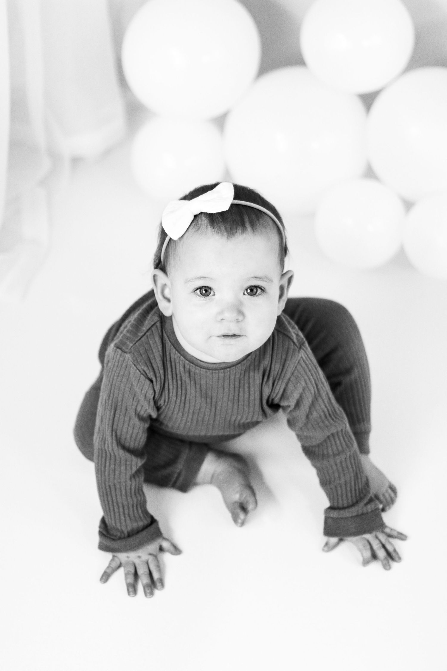 Black and white portrait of baby crawling with bow headband against white balloons background.