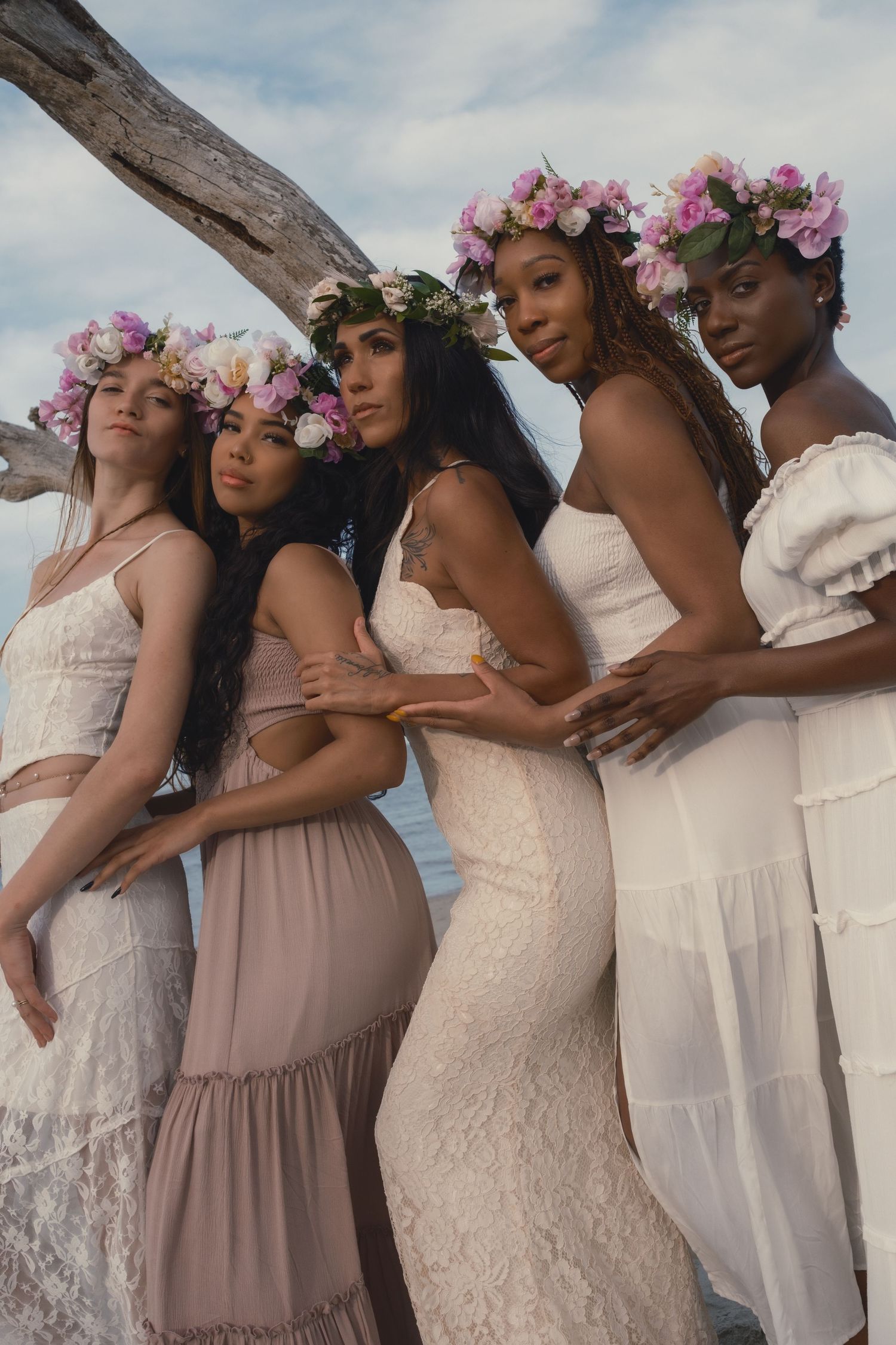 Group of women wearing flower crowns and flowing dresses standing together on a beach, radiating joy and unity.