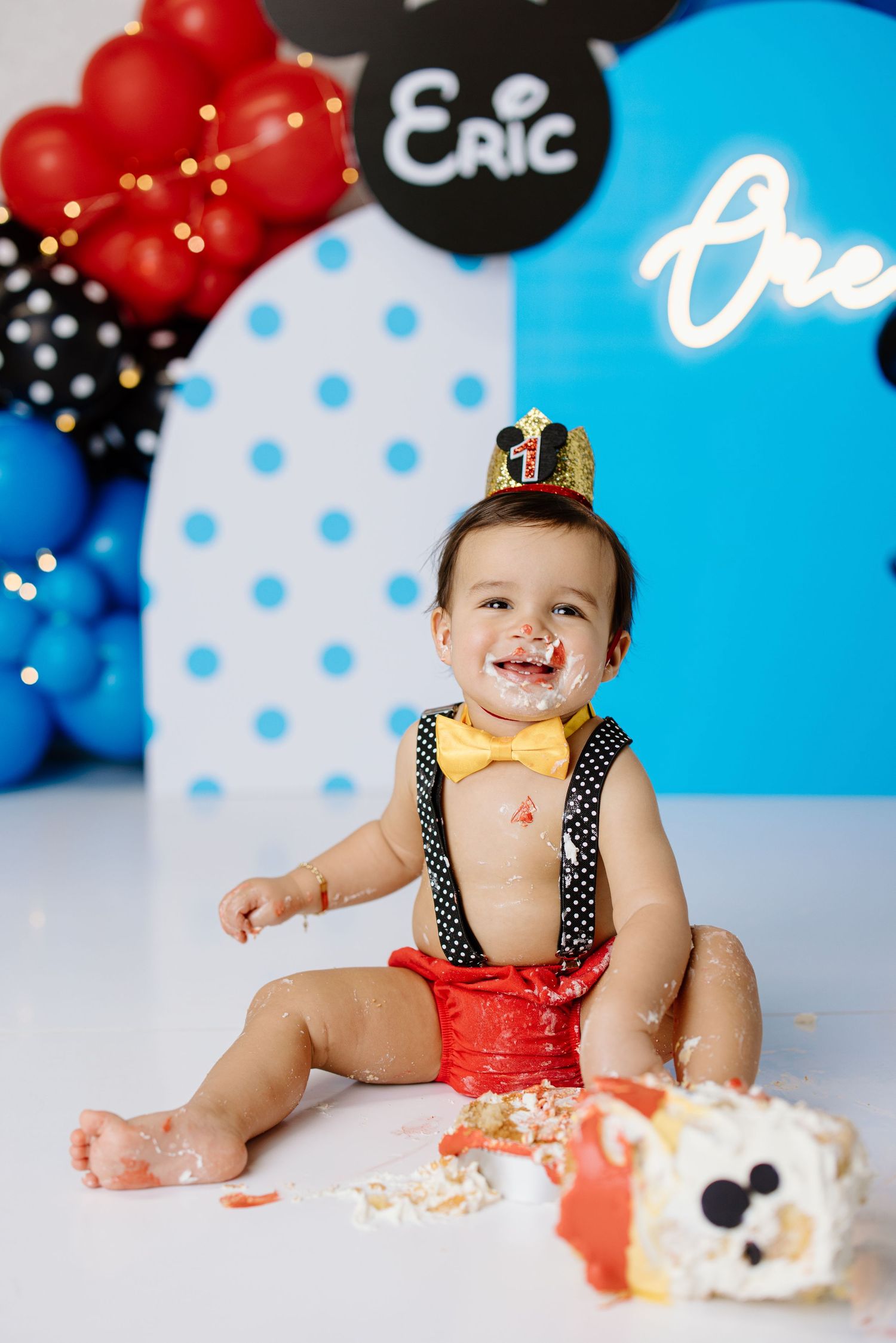 Festive first birthday cake smash setup with polka dot backdrop, balloons, and smashed cake on white floor.