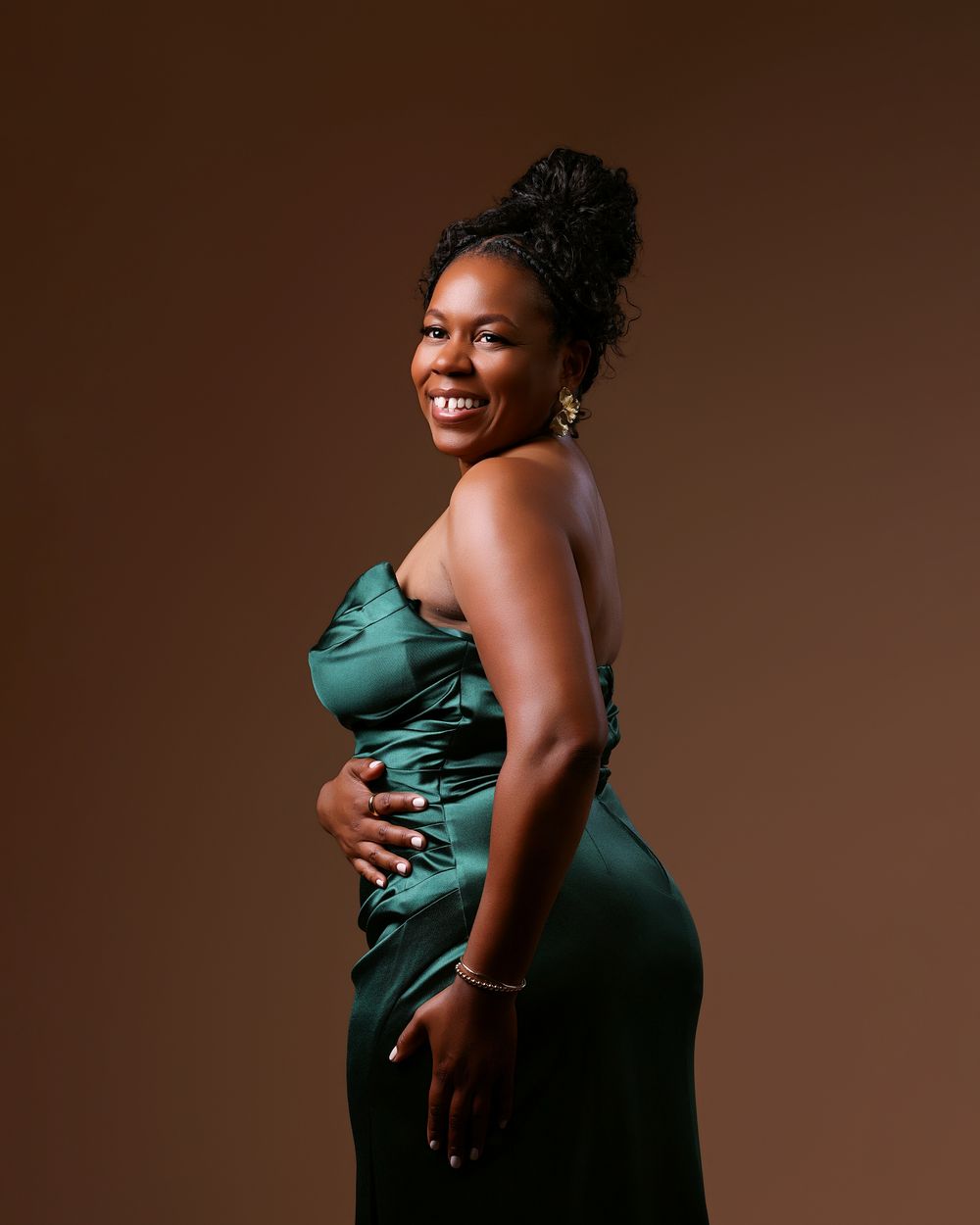 A smiling woman in an elegant emerald green satin gown poses confidently against a warm brown studio background.