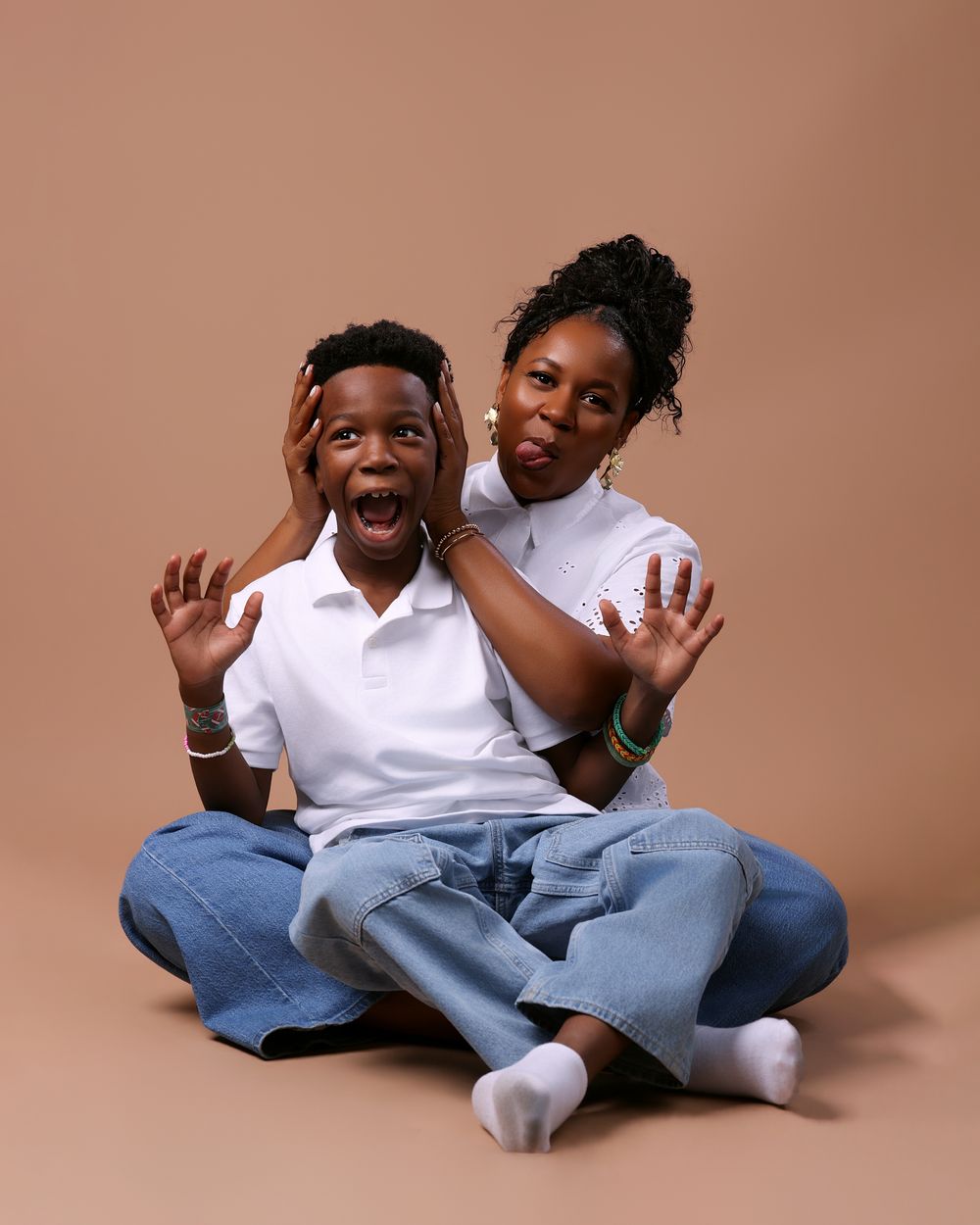 A woman playfully covers a young boy's ears as both make fun expressions, sitting against a beige background.