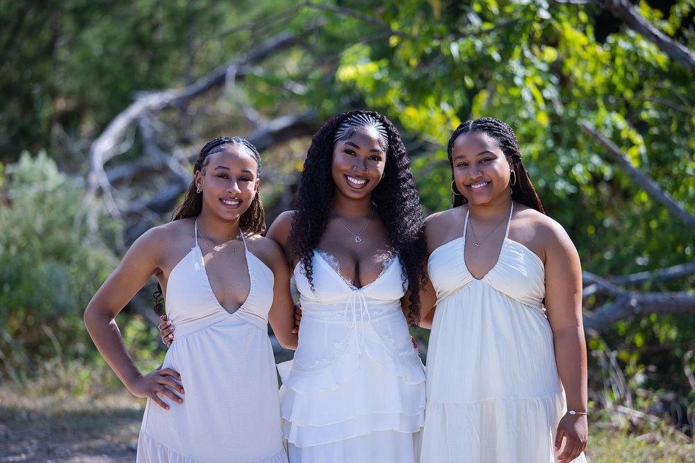 Three young women in white dresses smiling outdoors with green trees in the background on a sunny day.