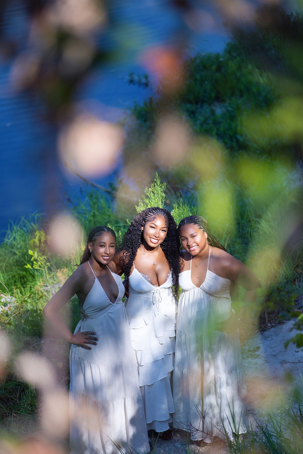 Three young women in white dresses pose together outdoors near water, framed by blurred foliage in the foreground.