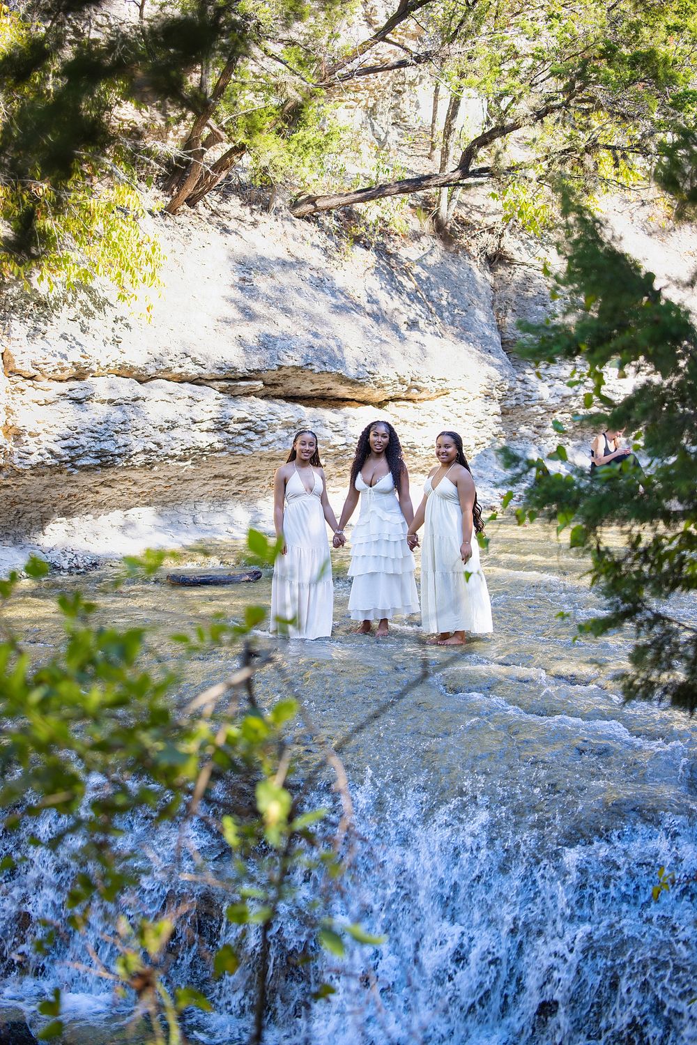 Three women in white dresses hold hands standing in a rocky stream surrounded by lush trees and a cascading waterfall.