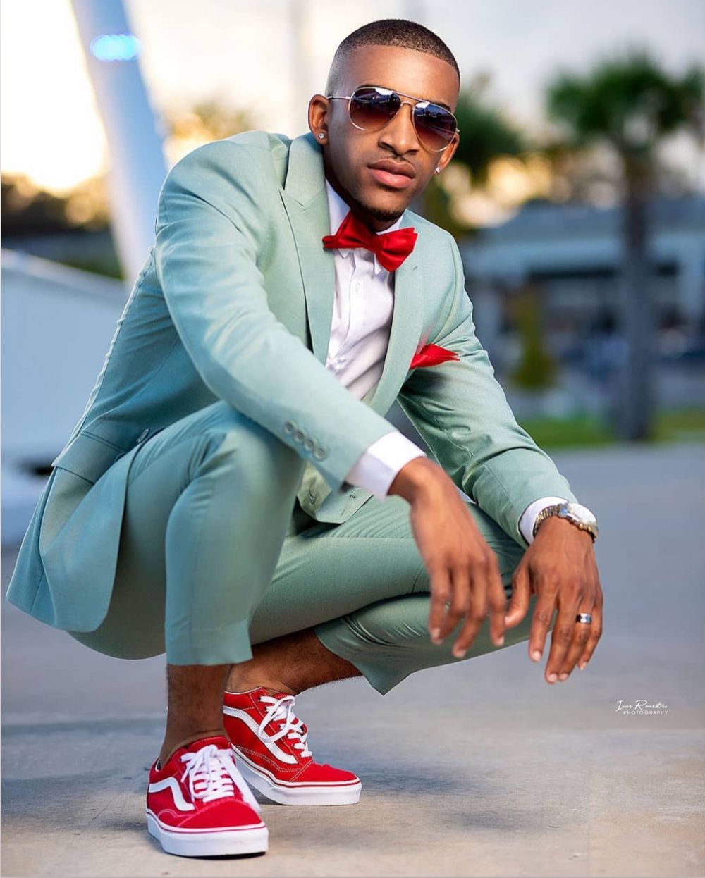 Stylish man in mint green suit with red bow tie and red Vans sneakers, crouching outdoors.