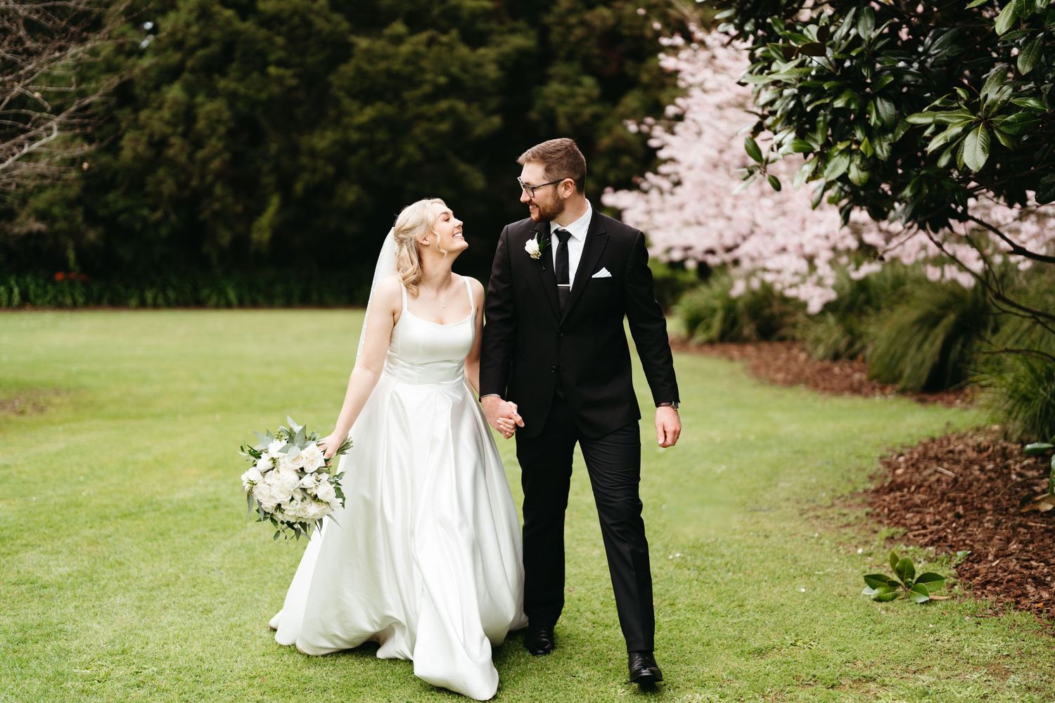A couple strolls through lush gardens in elegant wedding attire with white florals at Kumeu Valley Estate Wedding venue.