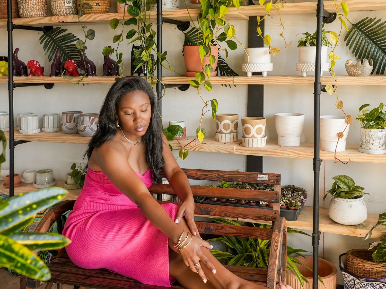 Woman in pink dress sits on bench surrounded by potted plants and wooden shelves.