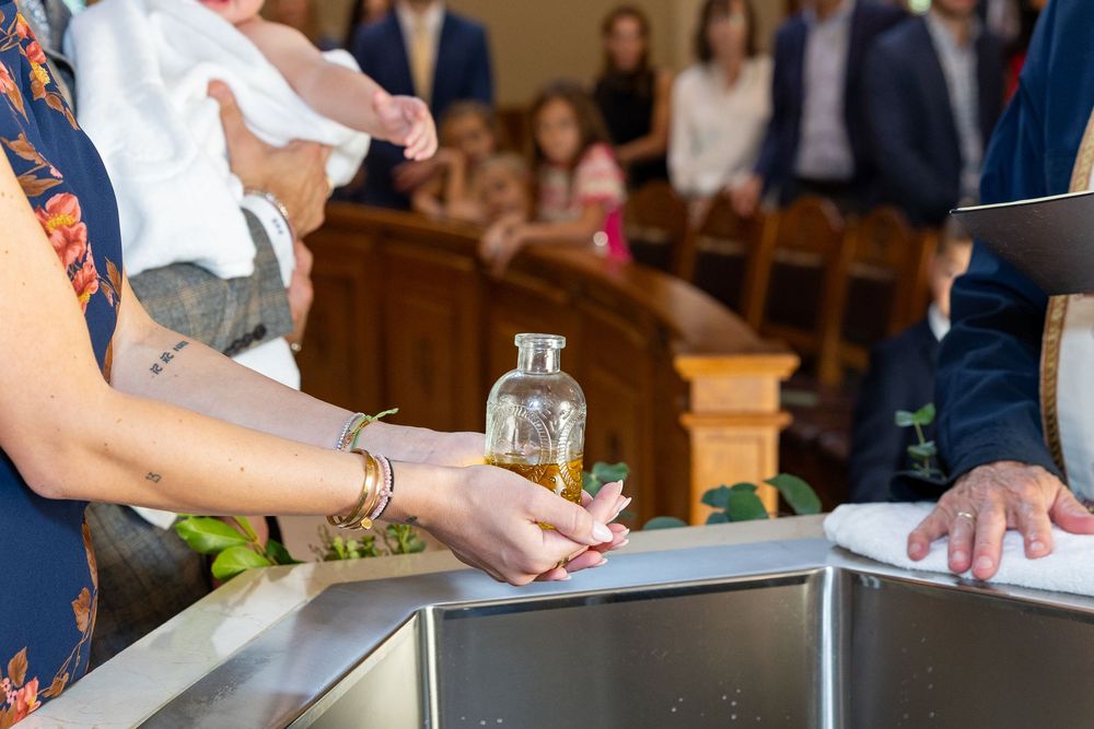 Godmother's hands hold a glass bottle with oil during a baptism ceremony at church with wooden pews visible in the background in Weston, MA.