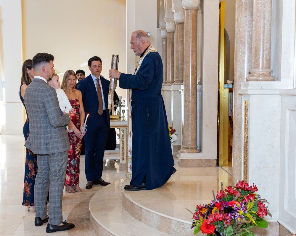 A family in formal attire gather in an elegant church with white columns and red floral arrangements during a baptism at St. Demetrios.
