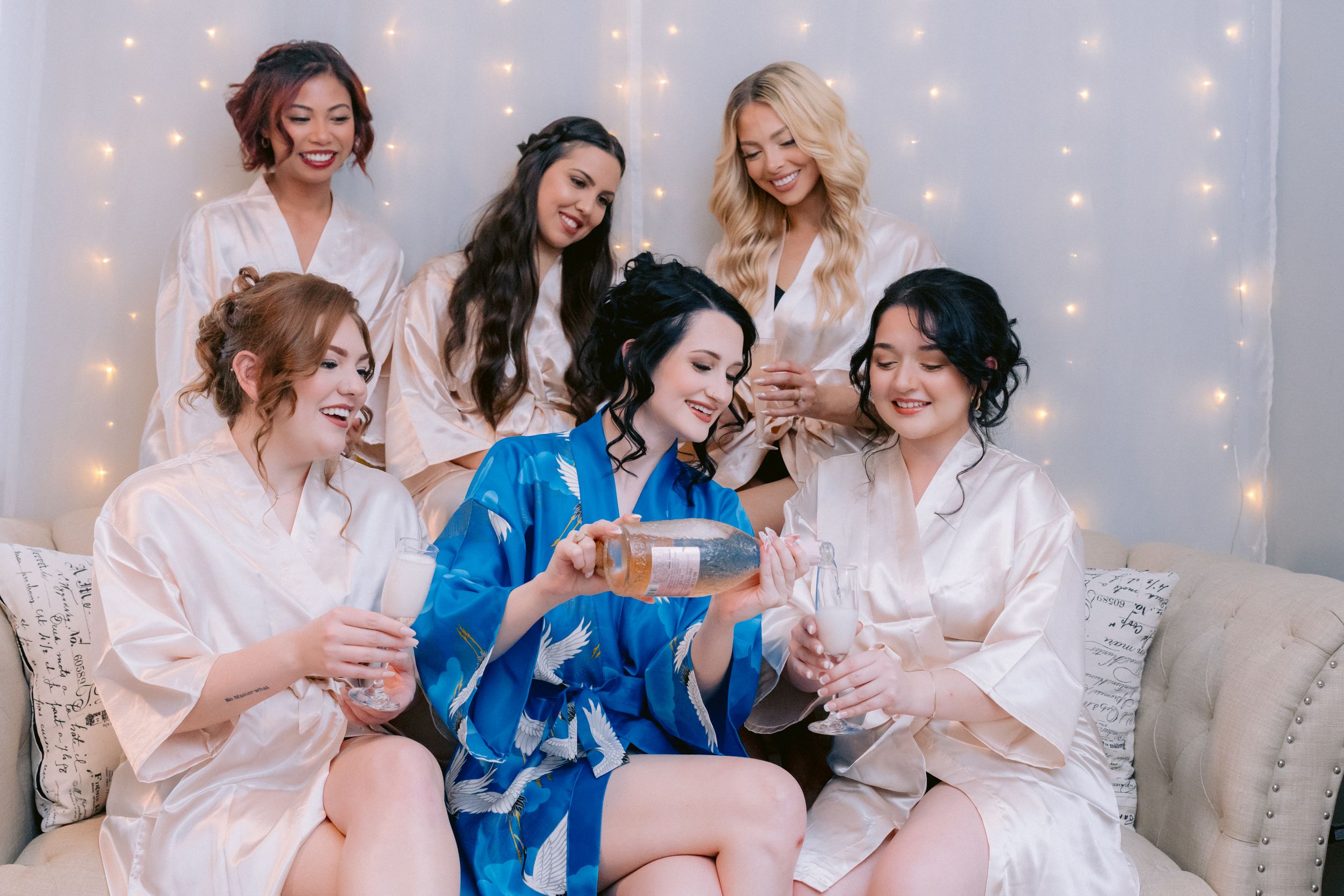 Friends in matching robes laugh together on a couch at a pre-wedding celebration with twinkle lights behind them.