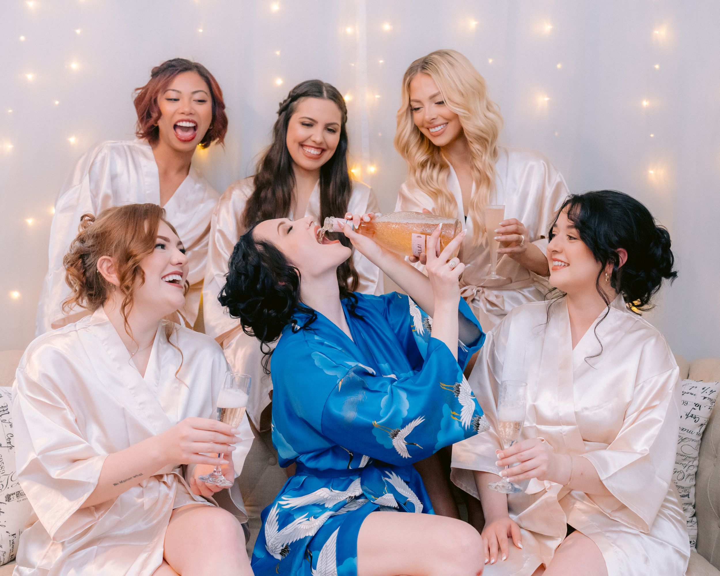 Group celebrates with champagne wearing matching silk robes against a backdrop of twinkling lights.