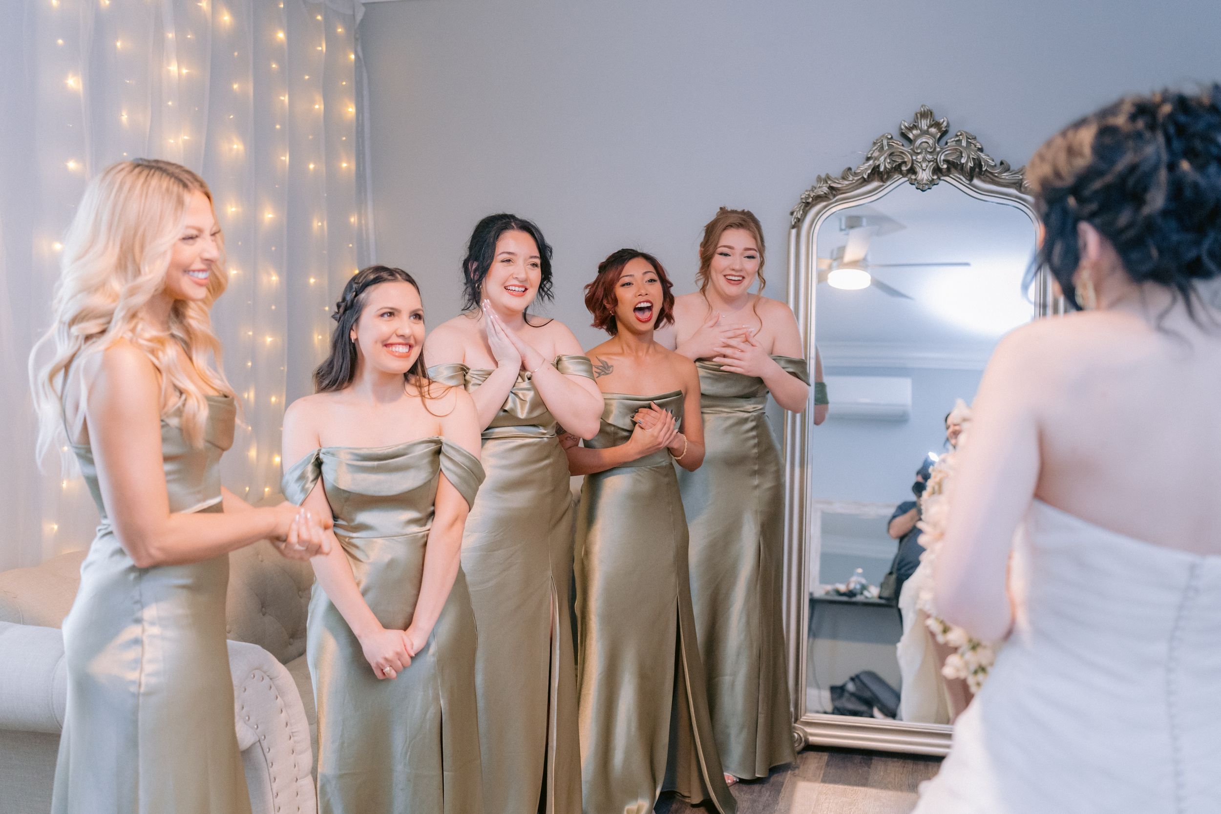 Wedding party in silk champagne dresses shares an excited moment in front of an ornate mirror.