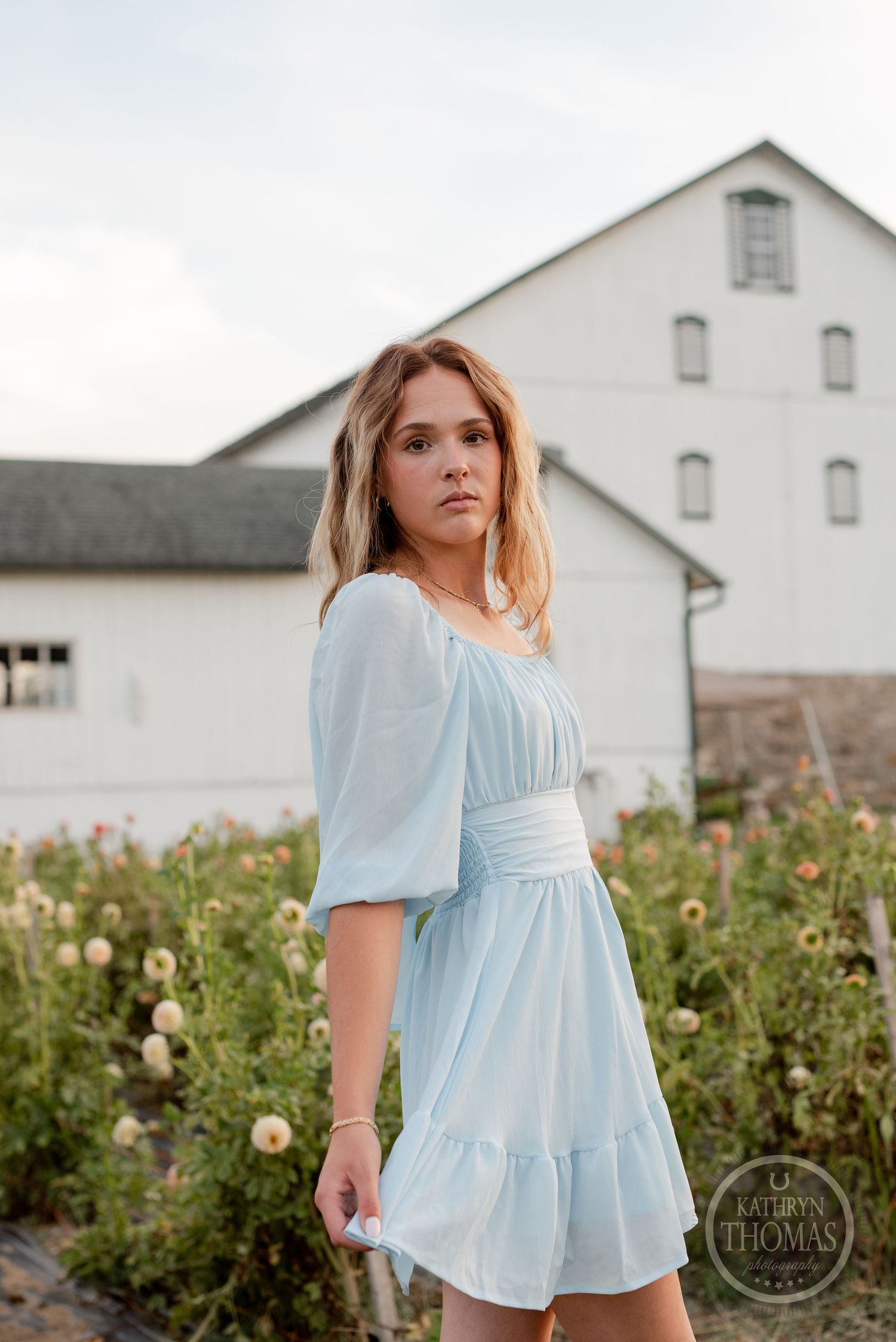 A delicate pale blue dress with puff sleeves stands out against a classic white barn and flower garden.