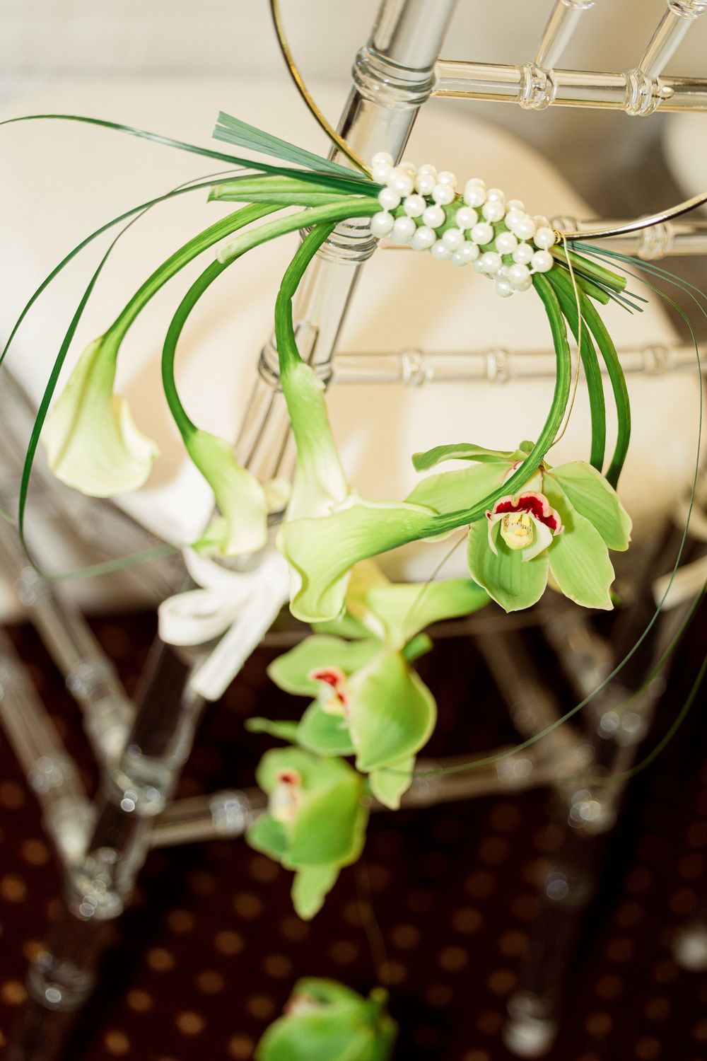 Elegant floral arrangement with green cymbidium orchids, white calla lilies, and pearl accents on a clear acrylic chair.