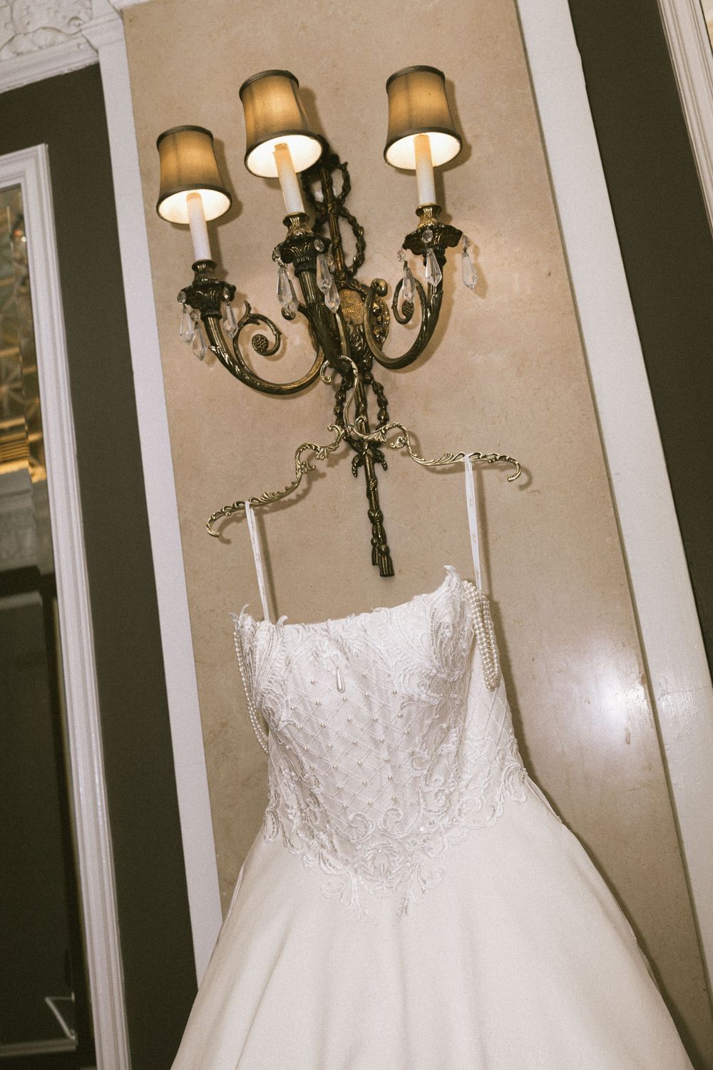White strapless wedding dress with lace bodice and flowing tulle skirt displayed against a wall with ornate lighting fixtures.