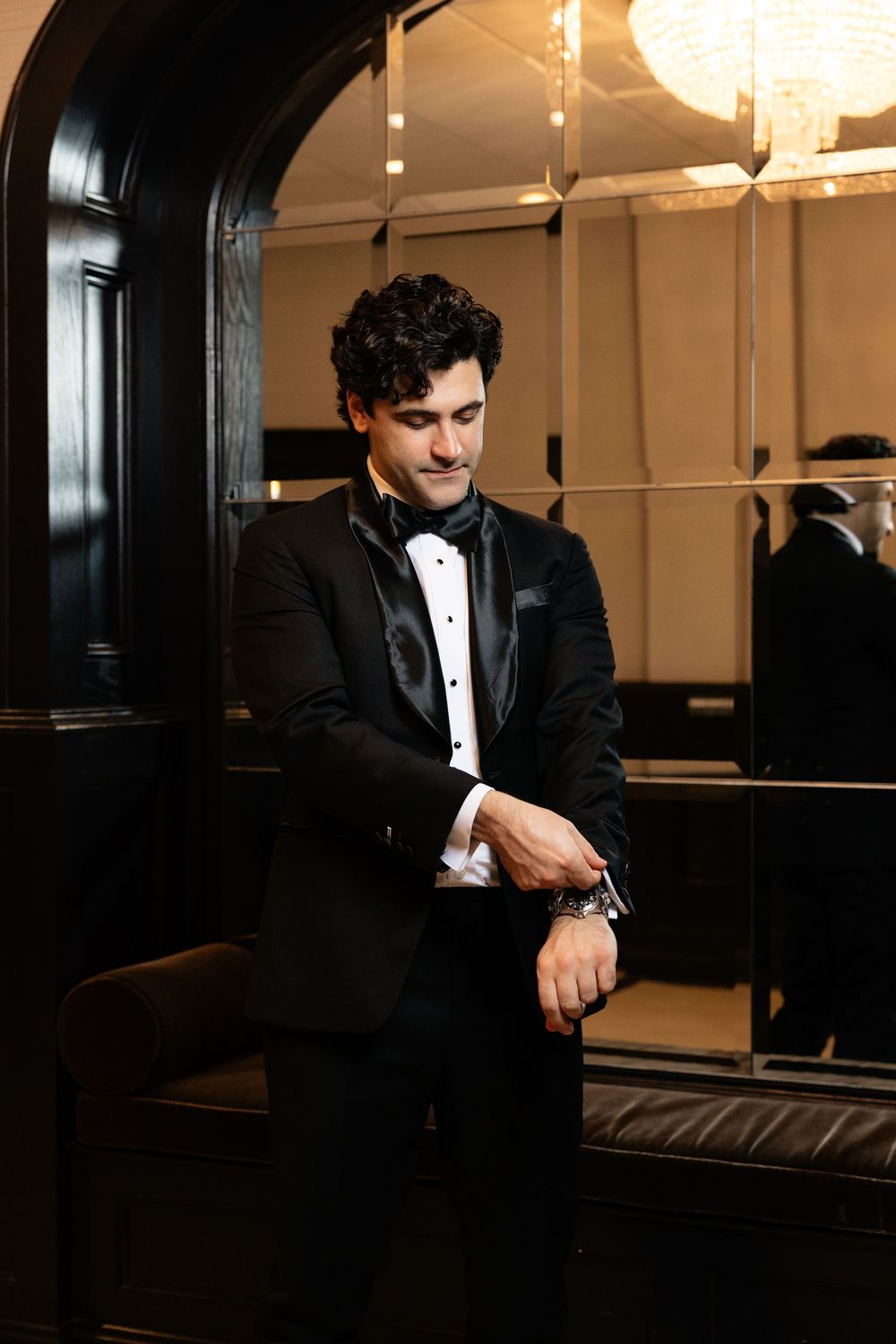 A person in a black tuxedo and bow tie stands in an elegant room with ornate lighting and dark wood paneling.