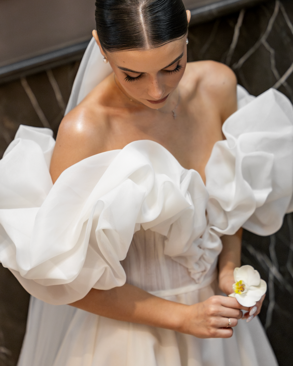 Elegant bride in off-shoulder wedding gown with ruffled details holds a delicate flower.