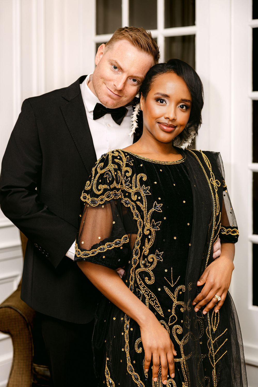 Elegant interracial couple posing in black formal attire, woman in gold-embroidered dress, man in tuxedo with bow tie.