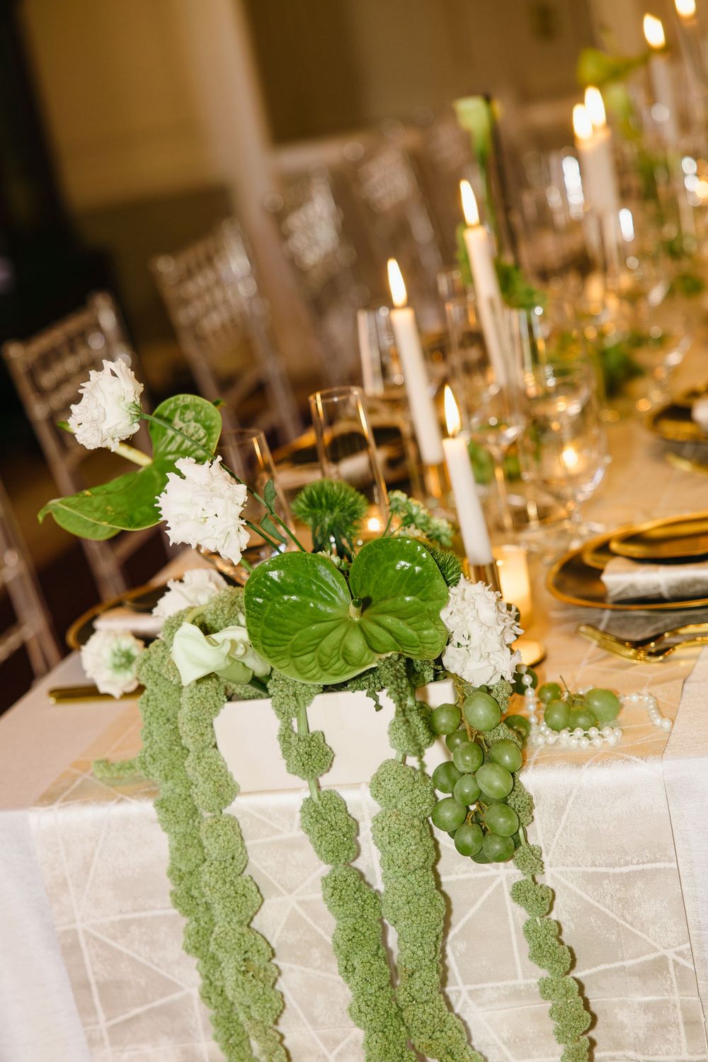 Elegant wedding table with green anthurium floral centerpiece, white candles, and gold accents on a white linen tablecloth.