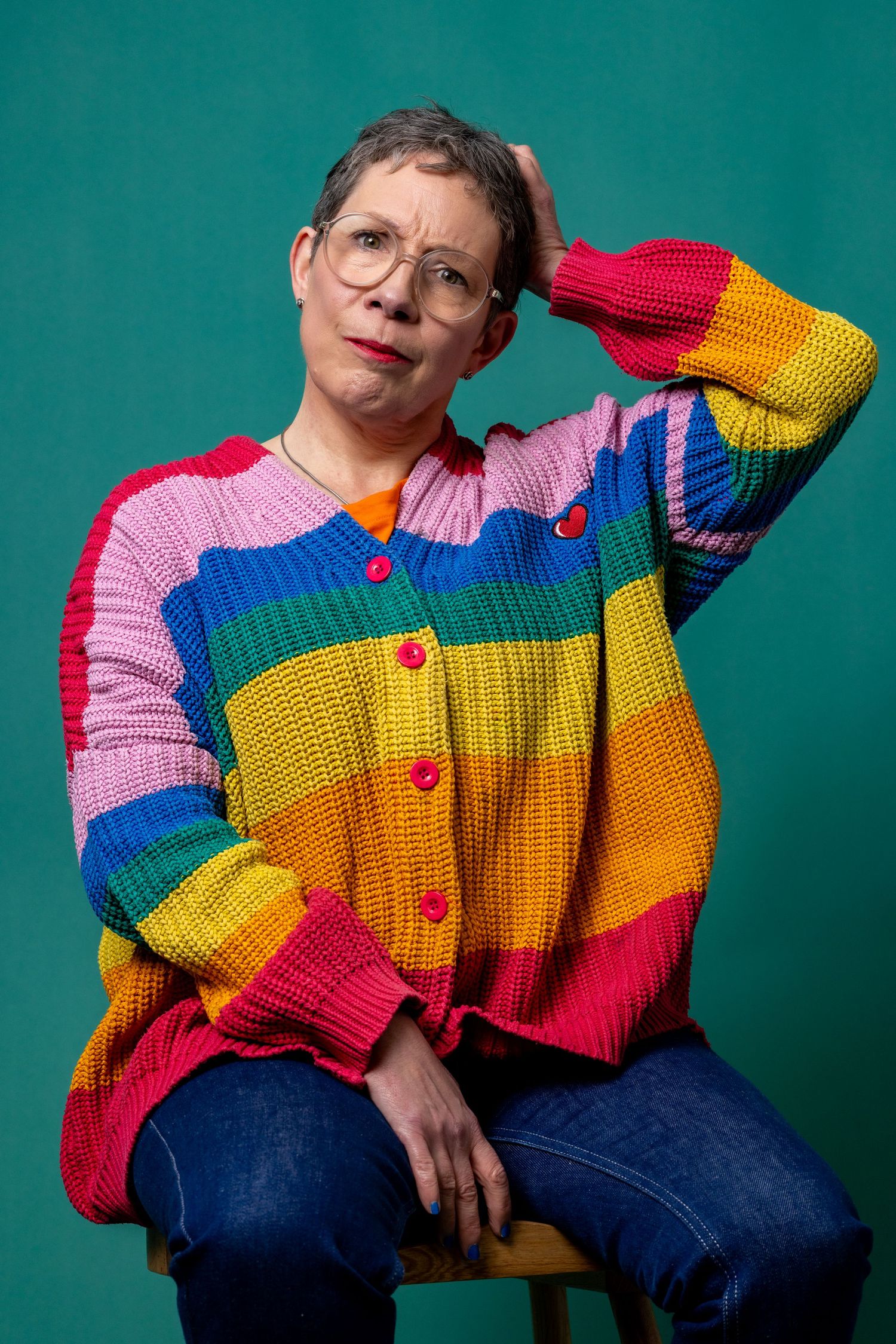 woman with short hair and glasses wearing a colorful, striped sweater, scratching her head with a puzzled expression