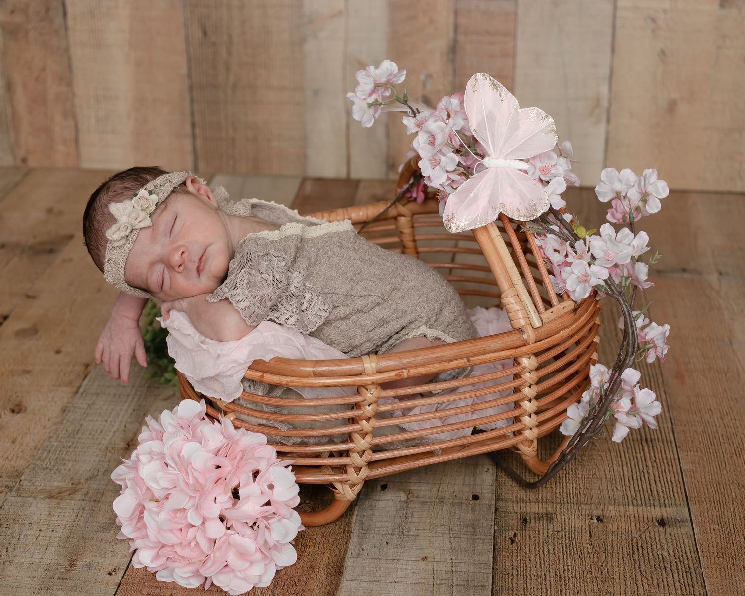 A sleeping newborn in a wicker basket surrounded by pink flowers and rustic wooden backdrop.