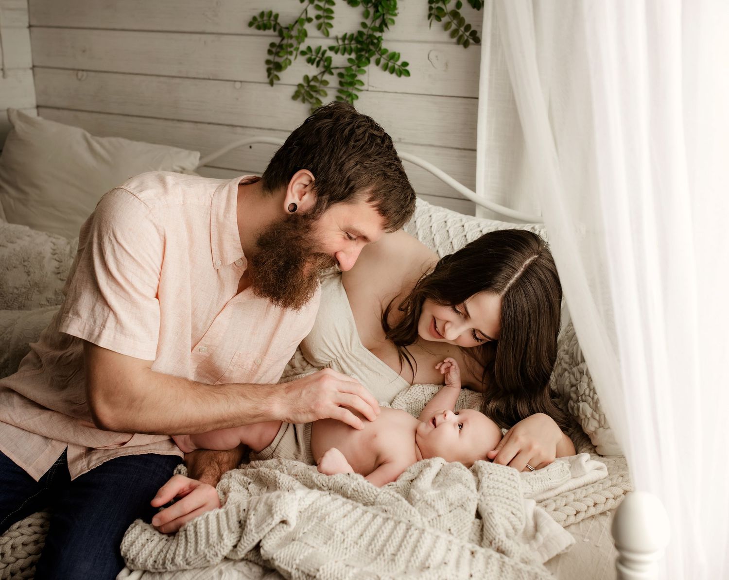 A bearded father and dark-haired mother lovingly gaze down at their newborn baby on a white knit blanket.