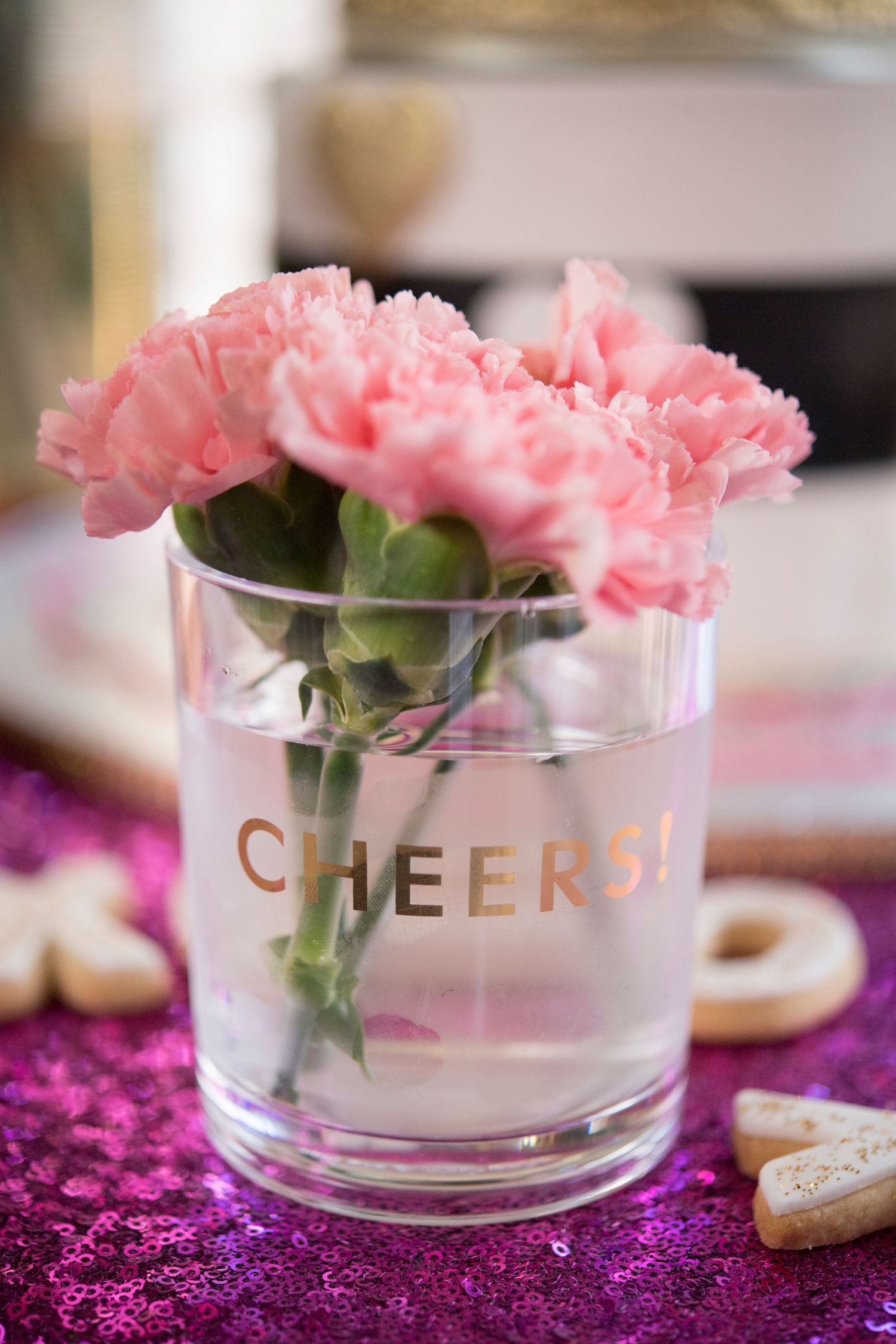 Pink carnations in a clear glass vase with 'CHEERS' text on sparkly pink tablecloth.