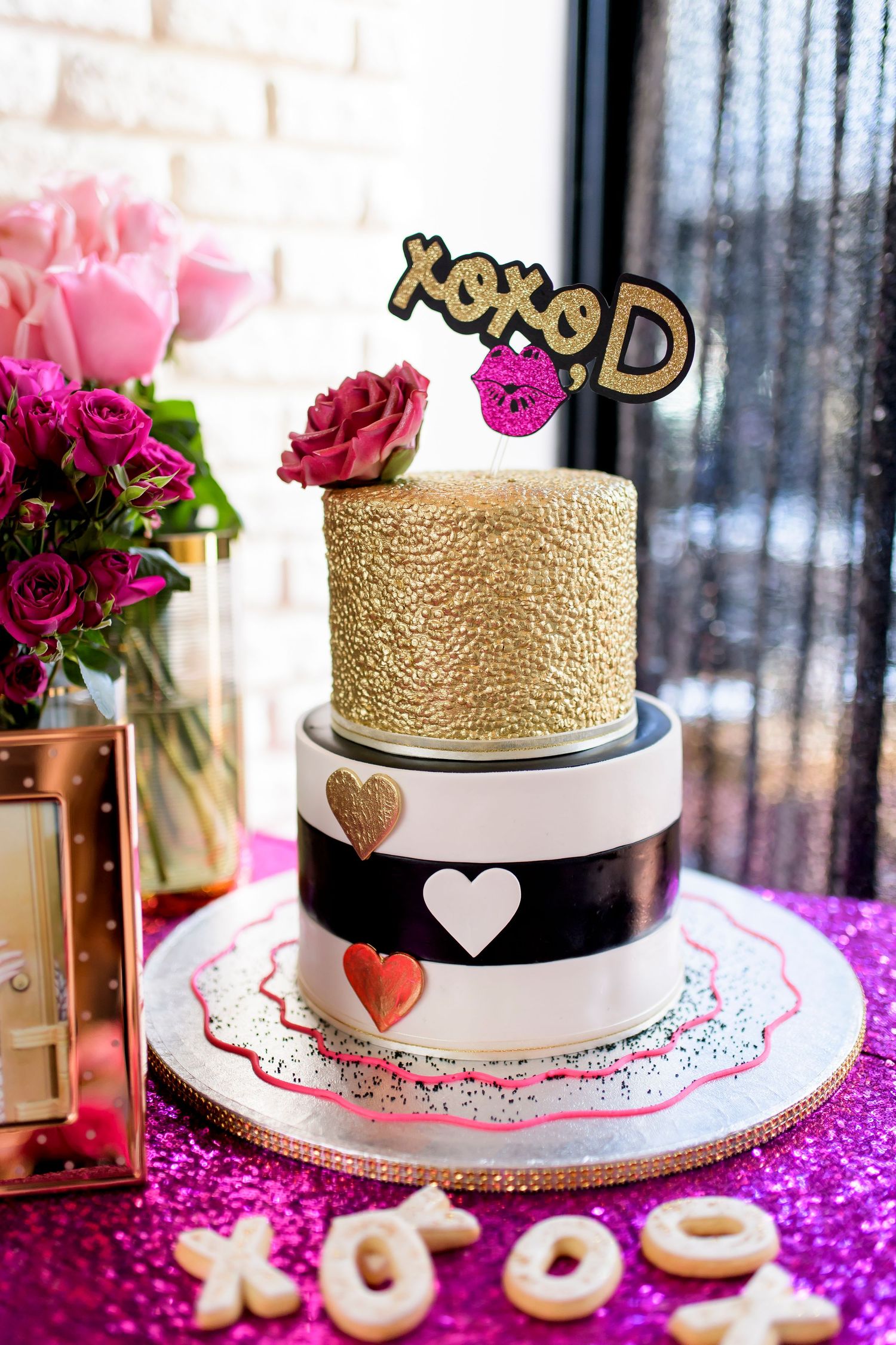 Three-tiered wedding cake with gold, white and black stripes topped with XOXO decorations and pink flowers.