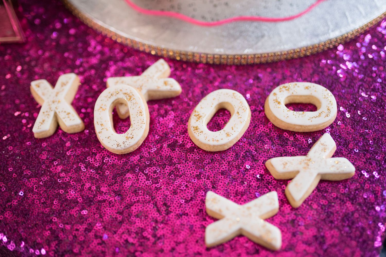 White XOXO letter cookies arranged on a sparkly pink sequin tablecloth.
